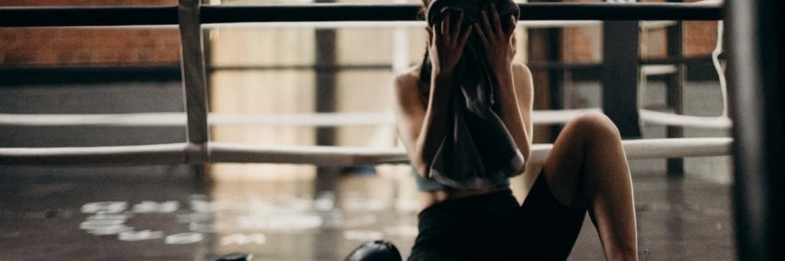 A female boxer sitting in a gym ring, resting and wiping sweat with a towel after a workout.